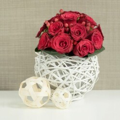 At the centre of the scene is a low, rounded floral centerpiece that feels purpose-made for an intimate wedding table or anniversary supper in Epping. A dome of luxuriant red roses dominates the frame, each bloom a deep, velvety crimson that almost seems to glow against the softer tones around it. The roses are arranged tightly together so their petals overlap in a continuous wave of colour, from tightly curled buds to fully open blossoms showing off their layered whorls. Slipped between the roses are polished clusters of red Hypericum berries, smaller and more compact, adding a berry-bright shine that catches the light and brings a gentle playfulness to the design. Just beneath the flowers, a discreet collar of dark green leaves peeks out, barely visible yet essential, offering a quiet contrast that makes the reds appear richer. The entire bouquet is cradled in a striking spherical vase formed from white-painted wicker or twigs, woven into an airy, open lattice. This sculptural white orb, perched on a pristine white tabletop, creates soft shadows and a sense of lightness that keeps the composition from feeling heavy. To the left, two decorative wicker spheres in a natural beige tone rest on the same surface, one large and one small, their woven strands echoing the texture of the vase while introducing a more rustic, earthy note. Behind, a softly blurred wall in gentle beige or taupe provides a calm backdrop, like the walls of a thoughtfully decorated Epping townhouse dining room or a local boutique venue. The whole image conveys warmth, romance, and contemporary elegance, ideal for envisioning how a refined red rose centerpiece could grace wedding receptions and special dinners across Epping and nearby villages.