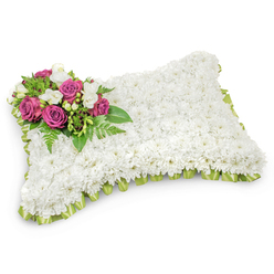 A lovingly constructed floral pillow composed of hundreds of small, immaculate white chrysanthemum heads creates a soft, cloud-like surface that speaks of peace and purity. The uniform white field is interrupted on the top-left by a poignant cluster of deep fuchsia and magenta roses whose rich tones convey affection and reverence; each rose's layered petals open gently as if cradling a memory. Nestled among them are sprigs of delicate white freesias or similar tiny blooms, their subtle fragrance suggesting tenderness and thoughtful remembrance. Verdant greenery, including feathery fern fronds and glossy foliage, provides a natural cradle for the accent group, framing the colour and giving depth to the design. Along the pillow's perimeter a pale green satin ribbon is neatly pleated, the sheen of the fabric catching soft light and lending a refined edge to the form. Photographed on a pristine white backdrop with a soft shadow beneath, the piece reads as both sculptural and intimate. As a local Epping florist, Flowers Epping often arranges this style for funerals and memorials along the High Street and for families gathering near Epping Forest or in nearby Theydon Bois; the design is chosen to offer quiet comfort when words are scarce. The composition balances texture, scent, and colour restraint-an understated but eloquent tribute crafted to honour a life and to provide solace in times of loss.