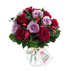 A dense, round bouquet composed of velvet-red and pale-lilac roses rests in an antique-look mercury glass vase, photographed against a stark white backdrop so every hue reads clearly. Plum and crimson roses form the heart of the arrangement, their petals layered and softly curled at the edges, intermingled with mauve and lavender blooms that breathe a cooler, serene note into the bouquet. Deep green foliage-the glossy, elongated leaves of what appears to be Ruscus or Salal-frames each bloom and gives the arrangement a grounded, garden-fresh structure. Tiny clusters of red-orange berries or rose hips are tucked between the roses; their firm, slightly glossy skins add a rustic counterpoint and a playful, seasonal accent. The vase is an important touch: rounded and slightly bulbous at the bottom, it narrows toward the top and is finished in a mottled, reflective mercury glass that picks up light in soft, antique highlights. Natural raffia or twine is wrapped around the neck, threaded through a simple white rectangle tag bearing a small hand-drawn rose motif and a cursive message in green-tinted ink. The overall impression is intimate and crafted-an arrangement that suggests quiet celebration, thoughtful condolence, or an anniversary gift. As a local florist might note, this is the kind of bouquet that travels well on short delivery routes across Epping town centre and toward the edge of Epping Forest, arriving smelling faintly of garden earth and summer evenings.