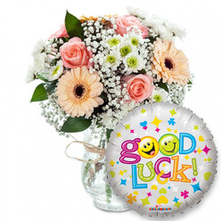 Framed on a crisp white background, this Good Luck gift pairing feels simultaneously handcrafted and celebratory - the kind of thoughtful composition we arrange at Flowers Epping for neighbours across Epping and along the High Street. To the left a full bouquet sits in a clear glass vase tied simply with rustic twine, the glass catching a little of the room's soft light. The flowers are a gentle chorus of pale pink and peach roses, their velvety petals close and reassuring, paired with peach-orange Gerbera daisies that offer sunny faces and dark brown centers for contrast. Wisps of baby's breath spill like soft lace between stems, while small white and lime-green chrysanthemum-like florets introduce a crisp, lively note. Rich green foliage frames the blooms, providing natural depth and a cooling undertone to the warm palette. In front and to the right, a round silver foil balloon reads Good Luck! in buoyant, multi-coloured letters, decorated with scattered hearts, stars and confetti shapes; the two o's in Good are replaced by bright yellow smiley faces - one winking, one cheekily sticking its tongue out - lending an affectionate, almost conspiratorial cheer. The overall effect is quietly joyful: scented roses and tactile petals balanced with the balloon's metallic sparkle, a bouquet and balloon meant to steady nerves, celebrate courage, or accompany someone setting off on a new path around Epping Forest or beyond.