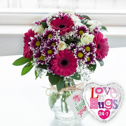 On a light, uncluttered surface that could easily be a kitchen table in an Epping townhouse, a vivid bouquet glows in gentle natural light. At its heart are bold gerbera daisies in saturated fuchsia-magenta, their smooth petals forming wide discs around dark, velvety centres that add dramatic depth. Interlaced throughout are neat clusters of chrysanthemums with eye-catching two-tone petals: deep purple at the base, fading to pristine white at the tips, each surrounding a sunshine-yellow centre that brings a sense of optimism. Between these, creamy white roses rest in varying stages of opening, their layered petals softening the design with a more traditional elegance. Fine sprays of white baby's breath appear to float between the larger blooms, creating a lace-like veil that softens edges and adds a gentle, romantic haze. Fresh green foliage emerges from beneath, shaping the bouquet into a rounded, balanced dome that feels thoughtfully composed. The clear glass vase reveals the straight, carefully trimmed stems immersed in clean water, with rustic natural twine tied around the neck in a relaxed bow. A petite white gift tag hangs from the twine, decorated with a pink rose emblem and friendly green handwriting that suggests the personal care of a local florist. To the right of the vase, a shiny heart-shaped foil balloon reflects the light, its silver-edged shape filled with playful, multi-coloured lettering that reads "Love n' Hugs 24-7," hinting at constant affection. The softly blurred, bright background evokes a window looking out toward Epping Forest or a quiet residential street, giving the entire scene a warm, homely feeling ideal for same-day flower delivery to friends, partners, or family nearby.