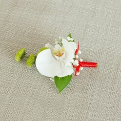 A single, pristine white Phalaenopsis orchid forms the serene centre of this boutonniere, laid carefully on a light beige, linen-like fabric whose subtle, slightly coarse weave adds a natural, understated backdrop. The orchid's petals are wide, smooth, and gently curved, catching soft, even light that highlights their porcelain-like texture. In the flower's throat, hints of buttery yellow and delicate reddish-orange markings create a warm focal point, adding character and depth to the bloom. Surrounding the orchid, airy sprays of baby's breath extend like a fine mist, their tiny white blossoms offering a cloud-soft contrast and a sense of lightness, as though the piece could almost float above the fabric. To one side, two bright lime-green chrysanthemum buds, round and intricately textured, introduce a crisp, contemporary note, their bold colour reminiscent of new leaves in the woodland edges around Epping Forest. A broad, deep green leaf cradles the orchid from beneath, while a smaller, darker leaf peeks out near the base, both adding structure and a lush, botanical richness. The stems are meticulously bound with a vivid crimson red satin ribbon, wrapped snugly to form a sleek handle, then finished with a tidy little knot or bow that injects a confident splash of colour. The composition feels calm yet celebratory, ideal for a groom or groomsman stepping into a church or registry office in Epping, the boutonniere acting as a quiet statement of style and care, crafted with the same attention to detail trusted by couples who turn to Flowers Epping for their wedding flowers.