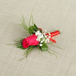 Set against a neutral, sand-beige fabric that looks like finely woven linen, a single-stem rose boutonnière becomes the quiet star of the image, as if waiting to be collected by a groom getting ready in an Epping townhouse near the station. The rosebud is a rich, velvety shade of deep fuchsia, edging toward crimson, its petals tightly wrapped so that the promise of full bloom is held close and intimate. Around this central rose, clusters of pure white baby's breath form a soft collar, each tiny floret with a gentle, cloud-like texture that contrasts beautifully with the rose's smooth, satin-like petals. Threading through and behind the blooms, slender strands of bright green, grass-like foliage and wispy fern fronds stretch outward, giving the boutonnière a fresh, just-picked-from-the-garden feel, the sort you might associate with early walks around the green spaces leading into Epping Forest. The stems at the base are carefully gathered and bound in a band of radiant red satin ribbon, meticulously wrapped so that it hugs the stems in a clean, seamless line, ending in a subtle loop that adds a touch of craftsmanship without distracting from the floral focal point. The lighting is bright yet soft, illuminating every vein in the leaves and every curve of the rosebud, making the colours feel true-to-life and celebratory. This handcrafted buttonhole conveys tenderness and ceremony, an ideal finishing detail for weddings, proms, or black-tie events across Epping, from intimate gatherings at local pubs to elegant receptions held along the High Street.
