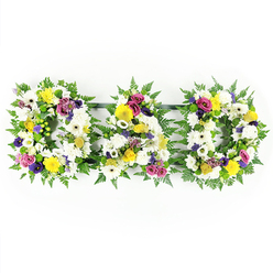 The image shows a deeply personal "DAD" funeral tribute, arranged as three separate block letters placed in a neat row on a brilliant white backdrop. Each sturdy letter is outlined with lush, arching fern fronds, their rich green tones framing the design and giving softness to the strong shapes. The faces of the letters are completely carpeted in white flowers - mainly large chrysanthemums and open gerbera daisies - so closely arranged that they form a continuous, cushion-like surface. Among this calm white canvas, little bursts of colour glow: cheerful yellow gerberas and small clusters of yellow spray roses add warmth, while lime-green button chrysanthemums and tiny pale green accents bring a fresh, springlike brightness. Scattered through the tribute, small groups of purple lisianthus and deep pink or magenta spray roses give depth and emotion, their jewel tones recalling love, respect, and cherished memories. The flowers all face outward, carefully graded in height so the letters read clearly even from a distance, as they might at a service in one of Epping's local chapels or at the cemetery on Bury Lane. Slim metal or wooden bars, hidden beneath the foliage, link the D, A, and final D, keeping the word steady and balanced as a single tribute. The overall effect is one of quiet strength and tenderness, like a final embrace expressed in flowers, thoughtfully designed by a florist who knows how families here in Epping wish to honour a much-loved dad with something that feels both personal and dignified.