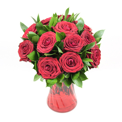 An opulent cluster of deep red roses fills a translucent red glass vase, the entire design photographed against a crisp, white background that lets every detail stand out with clarity. There are roughly a dozen to fifteen blooms, each rose carefully opened to that perfect stage where the petals have unfurled into generous spirals but still hold a sense of fullness and life, much like a bouquet you might have delivered to a townhouse near Epping Forest on a special evening. The petals are velvety and rich, forming concentric whorls that create a classic romantic rose shape, their intense crimson hue suggesting passion, romance, and enduring affection. Between the roses, substantial dark green foliage - likely ruscus or similar hardy greenery - provides a lush frame, each leaf smooth, slightly pointed, and gleaming as it catches the light. This foliage adds structure and volume, making the bouquet feel abundant and expertly balanced. The red glass vase that cradles the stems has a softly curved silhouette, narrowing towards the base and flaring gently at the top. Its subtly ribbed or wavy texture plays with light and shadow, introducing movement and nuance without distracting from the flowers themselves. Through the glass, hints of stems and variations in red deepen the sense of cohesion. The overall impression is of a carefully crafted, premium arrangement that would be chosen for heartfelt moments - perhaps a surprise anniversary delivery to a home off Station Road or a romantic gesture sent across Epping by a local florist who understands how powerful a dozen red roses can be when words are not quite enough.