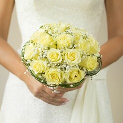Bathed in the intimate glow of an Epping afternoon, this expertly crafted bridal bouquet rests in the hands of a bride ready to step onto the leafy paths near Epping Forest. Soft yellow roses, perfectly shaped and delicately layered, are gathered in a tight, voluminous dome. Each petal is radiant, edged in pale gold and buttercream, giving the arrangement an inviting luminosity. Scattered throughout, dainty white tufts of gypsophila-known more fondly as baby's breath-add a dreamy lightness reminiscent of the early morning mists that drift through local parks. The entire composition is encircled at the base by looping bands of dark green grass, their subtle structure echoing the careful symmetry of expert floristry and a touch of Epping's rural elegance. The bride's gracefully manicured hands, nails blush-tinted and adorned with a slender ring, embrace the bouquet with care and pride. Soft lace textures of the wedding dress, perhaps inspired by the heritage of Epping's traditional styles, provide a serene, tactile backdrop. The background, softly blurred, draws every gaze toward the bouquet, as if pausing time on a significant local celebration. The mood is tender, suffused with hope and quiet joy, ideal for marking life's most cherished moments and for couples embarking on their next chapter in Epping's welcoming community.