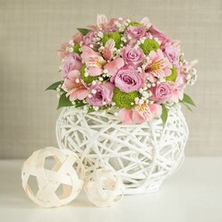 Imagine stepping into a sunlit Epping dining room-here, a lush centerpiece commands the scene, seamlessly balancing traditional romance with modern artistry. Atop a flawless white surface, the bouquet's pastel pink and lavender roses offer the gentle confidence of full bloom, surrounded by vibrant lime green chrysanthemums for a cheerful twist. Intertwined among the focal flowers are graceful sprays of baby's breath, their delicate white blossoms creating a sensation of openness and ease, much like the fresh air on a spring walk through Epping's ancient woodland. Pink alstroemeria lilies, with their trademark stripes and soft yellow accents, lend intricate detail and a light, welcome fragrance. The flowers unfold from a distinctive white vase: an openwork marvel crafted from fine, interlacing strips, both sturdy and stylish-its rounded silhouette and airy lattice design bring to mind the blend of rural charm and contemporary living present in homes near Epping's central green. Set beside the vase, two woven decorative spheres invite the eye with their natural-toned, basket-like texture and simple forms, each echoing the vase in miniature. Together, these elements create a scene that is both soothing and inviting. The overall effect is one of assurance and delight, perfect for elevating celebrations, weddings, or home gatherings in Epping, where fresh flowers from the local market always find their place among cherished décor.