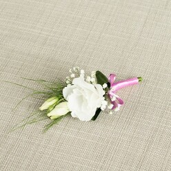 A single, fully opened white lisianthus bloom forms the graceful centre of this wedding boutonnière, resting at a slight angle on a pale beige, linen-like fabric that recalls the soft, natural textures often seen in Epping's country-style venues. The lisianthus petals are gently ruffled, layered in soft curves that create a rounded, almost rose-like form, their crisp whiteness glowing subtly in the even, diffused light. Just below and to the left of the main flower, two elongated lisianthus buds, still closed and tinged with creamy green, lean together like a quiet echo of the central blossom, suggesting new beginnings and the unfolding of a shared future. Fine sprays of white gypsophila thread through the design, their tiny, cloud-like flowers adding delicacy, volume, and a feeling of airiness, much like light mist over the paths of nearby Epping Forest in early morning. Slender, grass-like stems of greenery stretch outward in graceful arcs, adding gentle movement and texture, while a deeper, broader leaf tucked just behind the lisianthus helps anchor the arrangement with its rich, velvety green. At the base, the florist has bound the stems with a vivid fuchsia satin ribbon, tied into a neat, perky bow that introduces a confident splash of colour and a slightly playful note to the otherwise classic white-and-green palette. The ribbon's silky sheen catches the light and draws the eye, making this boutonnière ideal for a groom, groomsman, or prom guest who wants a subtle but memorable floral accent. The whole piece feels fresh, carefully composed, and perfectly suited to elegant celebrations across Epping and the surrounding Essex villages.