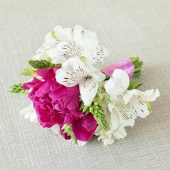 A beautifully crafted small bouquet, likely intended as a boutonnière or delicate wrist corsage, rests gently on a pale beige, linen-textured cloth that gives the image a calm, natural backdrop. The eye is immediately drawn to a single, fully open peony-like bloom in a vivid fuchsia-magenta tone, its densely layered petals ruffled and slightly cupped, forming a plush, cushiony centre that suggests softness and fullness. Surrounding this striking flower are several white alstroemeria, or Peruvian lilies, each with six slender petals edged in pure white and marked inside with fine, burgundy and maroon streaks that radiate from the throat like elegant handwriting. Threaded between the larger blossoms are slim spikes of fresh green buds and narrow, elongated florets, their bright, almost lime-toned green foliage bringing a lively freshness and gentle movement to the composition. All of the stems are carefully bundled together and finished with a narrow band of bright pink satin ribbon, tied smoothly and snugly so that its glossy sheen mirrors the depth of the central bloom's colour. Soft, diffused lighting creates a feeling of calm, with gentle highlights gliding over the petals and subtle shadows nestling into the folds, evoking the quiet anticipation of a wedding morning in Epping. It feels like a piece a local florist might prepare for a ceremony near Epping Forest or a celebration at a nearby country pub, radiating romance, joy, and a sense of personal detail that will be remembered long after the petals fade.