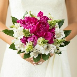 A detailed close-up shows a round, hand-tied bridal bouquet held just in front of a white wedding dress, the lace and fabric blurred softly so the flowers take centre stage. Dominating the arrangement are opulent peonies in saturated fuchsia and magenta, their petals layered and ruffled like fine silk, creating a plush, almost cloud-like heart to the bouquet. Slender stems of white alstroemeria weave between them, each trumpet-shaped flower marked with intricate maroon speckles and fine lines along the inner petals, bringing an elegant, slightly exotic character to the design. Threaded through this mix are bright green, spiky buds and textural elements, likely Ornithogalum or a similar variety, their pointed forms and crisp colour injecting energy and movement, much like the fresh green you see around the edges of Epping Forest in early summer. A collar of deep green, glossy foliage frames the flowers, giving the bouquet a polished outline while still feeling natural and garden-inspired. The bride's hands, visible at the base with soft pink polish on neatly shaped nails, gently enclose the stems, hinting at a moment of calm anticipation before the ceremony. The lighting is even and flattering, highlighting every fold of petal and every shift of colour from deep magenta to creamy white and vibrant green. Crafted in a style that brides across Epping often request for romantic, modern weddings, this bouquet balances bold colour with classic shape, making it ideal for capturing those close-up photos outside local venues and for inspiring couples planning their own wedding flowers in the area.