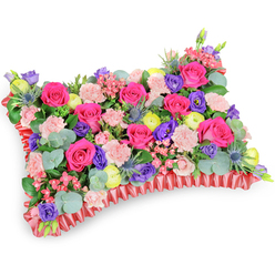 Seen from a gentle aerial angle, this rectangular sympathy cushion is a masterclass in texture and colour balance, an arrangement I'd prepare personally at Flowers Epping for families gathering near Epping High Street or beneath the oaks of Epping Forest. The surface of the pillow is densely packed with hot pink roses-some tightly furled, others opening to reveal a velvety heart-interlaced with soft, pale pink carnations that create a ruffled, cloud-like softness. Purple lisianthus adds depth with its graceful bells while bright lemon-yellow ranunculus speckle the composition, providing sunny accents that suggest warmth and celebration of life. Silver-blue eryngium thistles bring an unexpected architectural edge, their spiky forms punctuating the softer blooms and lending subtle cool tones against the warm palette. Rounds of silvery eucalyptus and lush green foliage are threaded throughout, providing structure, scent and a cooling contrast to the vivid flowers. The arrangement rests on a deep red, pleated satin border-the ruffled ribbon base frames the cushion and gives a dignified finish suited to memorial settings. The lighting is soft and even, picking up the gentle sheen of petals and the satin ruffle while preserving a sense of calm. Designed to offer comfort more than sorrow, this floral pillow reads as an affectionate tribute-vivid fuchsia, violet, lemon and blush harmonise to honour memory, suitable for funeral services, memorial tables, or as a condolence gesture delivered locally across Epping and surrounding villages.