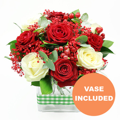 In this detailed floral image, a luxurious bouquet of red and white roses is presented front and centre, crafted with the kind of care you might expect from a local Epping florist who loves working with seasonal colours. The arrangement is dominated by plush, deep red roses and soft, creamy white roses, each flower fully open, with their layered petals forming graceful, cup-shaped blooms. Around them, clusters of tiny, vivid red filler flowers-similar to coloured gypsophila-create a fine, airy mist of colour, adding lightness to the dense rose heads. Interspersed red hypericum berries stand out as smooth, rounded beads, their lacquered surfaces hinting at winter berries found along paths near Epping Forest. Lush greenery anchors the display, with eucalyptus leaves and other mixed foliage adding tones of emerald and muted blue-green, giving a modern twist to a classic colour palette. The bouquet is arranged in a clear, rectangular glass vase, through which the neatly trimmed stems are visible, emphasising the tidy, professional finish. A green and white checkered gingham ribbon encircles the middle of the vase like a cheerful sash, suggesting a cosy, country kitchen or a festive table in a home near Epping High Street or Bell Common. In the lower right corner of the photograph, an orange circular graphic declares "VASE INCLUDED" in bold white capitals, making it clear that this floral gift arrives ready to place on a sideboard, office reception, or dining table. The overall mood is celebratory yet refined, perfect for anniversaries, winter gatherings, or a romantic surprise delivered across the Epping area.
