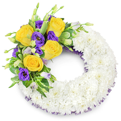 There is a calm, deliberate artistry in this circular sympathy wreath, shaped like a reassuring ring and intended to offer comfort in a time of loss in Epping. The bulk of the piece is a continuous band of fresh white spray chrysanthemums, each tiny bloom closely placed to create a dense, cloud-like cushion of petals; the uniform white surface reads as purity and quiet respect. Nestled into the upper-left quadrant, a cluster of accent blooms brings gentle colour and sentiment: warm, open yellow roses whose petals spiral softly outward and suggest a memory held tenderly, paired with deep purple lisianthus whose ruffled, satiny cups introduce a cool, dignified contrast. Sprigs of unopened lisianthus buds in softer lavender and tips of green add visual depth and the sense of life continuing. Varied greenery - rounded glossy leaves, some with faint variegation, and slim fern fronds - weaves through the spray to support and separate the focal flowers, creating movement and natural balance. A thin, dark-purple mesh ribbon slips beneath the chrysanthemums around the ring's edges, a subtle detail that ties the whole colour story together. Set against a stark white backdrop, the wreath's open centre and meticulous construction emphasize its traditional purpose as a funeral tribute: solemn, compassionate, and hand-finished. As a florist I picture this piece placed beside a church lectern or carried into a chapel near Epping High Street - a considered expression of condolence that feels both respectful and quietly warm.