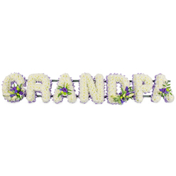 The image shows a carefully handcrafted funeral flower tribute that spells GRANDPA in bold, upright capital letters, stretching across the frame like a gentle banner of remembrance. Each letter is fully carpeted with compact white chrysanthemums or carnations, chosen for their purity and lasting freshness, the blossoms pressed closely together so the surface appears soft, almost velvety. The white blooms catch the light evenly, creating a serene, luminous effect against the plain background, much like a still moment of reflection in Stonards Hill or one of Epping's quiet residential lanes. Threaded through this sea of white are delicate accents of purple and lavender flowers with touches of bright green foliage, thoughtfully arranged to sit within the curves of the G, the spaces of the As, along the upright of the D, and rounding the bowl of the P. These small clusters resemble miniature posies, adding depth and a tender hint of colour, as if symbolising special memories or shared smiles. A fine edging of violet ribbon traces the outer contours of each letter, neatly outlining their shapes and giving the tribute a composed, finished appearance. Beneath the flowers, a sturdy dark green support bar quietly links the letters together, ready to rest beside a coffin or at the front of a chapel during a farewell service in Epping or the surrounding villages. The arrangement feels deeply personal, crafted with the sort of attentive care a local florist would give when helping a family honour their grandfather's life and the years of guidance, humour, and warmth he offered.