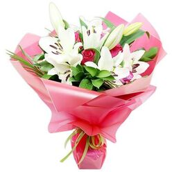 This image shows an exquisitely crafted bouquet that captures the romance of white Oriental lilies paired with deep red roses, presented in a way that feels both modern and timeless. The lilies dominate the composition, their pristine white petals sweeping outward in elegant arcs, each bloom centred by bold, dark stamens dusted with pollen, creating a striking contrast against the softness of the petals. Some flowers are fully open, revealing their trumpet-like forms, while others are still held in tapered buds, tinged with pale green and cream, suggesting an arrangement created fresh that morning in an Epping florist's studio. Between the lilies, clusters of red rosebuds emerge like jewels, their tightly coiled petals rich and velvety, with a few blossoms just beginning to unfurl to reveal darker inner tones. Deep green leaves and slender blades of foliage weave through the design, giving the bouquet height, structure, and a sense of natural abundance, as if gathered from gardens around Epping and the fringes of Epping Forest. The stems are enveloped in a voluminous layer of bright pink, glossy wrapping, slightly translucent so that hints of the greenery beneath show through, its rippling folds catching the light and adding a celebratory sheen. At the base, a slim lime green ribbon is tied neatly, its fresh colour lifting the palette and echoing the foliage. The overall impression is of a joyful, heartfelt gift, ideal for same-day delivery across Epping, Loughton, and nearby villages, perfect for anniversaries, Mother's Day, or simply sending love when words alone are not enough.