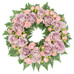 A serene, circular floral wreath is presented flat against a bright white backdrop, the overhead view capturing the quiet balance of colour and texture in this funeral tribute. The design forms a perfect ring, closely woven with large roses in muted shades of dusty lavender, soft mauve, and grey-tinged purple, each rose fully open and velvety, their ruffled petals creating gentle spirals that draw the eye around the circle. Nestled between these main blooms are clusters of smaller blush-pink spray roses, from tiny unopened buds to half-open flowers, which add a tender, almost whispering charm and a sense of gradual unfolding. The flowers sit atop a dense collar of deep green fern fronds, likely leatherleaf and similar textured greenery, whose finely divided leaves stretch outward to create a lush border that cradles the pastel tones at the wreath's heart. Small touches of darker foliage and miniature buds are tucked into the gaps, lending a soft shadow and depth that makes the roses appear even more prominent and alive. The arrangement feels quietly luxurious yet restrained, the sort of wreath chosen to convey sympathy, remembrance, and peace at a funeral or celebration of life gathering in Epping, perhaps near the forest edge or at a local chapel. Designed with care, the colours suggest enduring affection and gentle farewell rather than stark sorrow, offering comfort to families and friends. The fresh blooms and meticulous craftsmanship reflect the steady, thoughtful approach of Epping florists who understand how flowers can speak with dignity when words are hard to find.