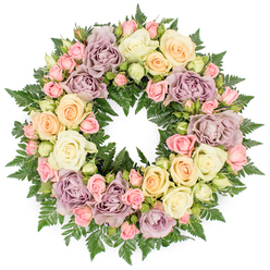 A classic circular memorial wreath is pictured against a clean, white studio background, its generous ring of roses and ferns forming a seamless halo of soft colour. The design is lush and tightly arranged, with no bare spaces, suggesting the careful, steady hands of an experienced Epping florist who understands the importance of every detail for a funeral or remembrance service. Large roses in creamy white and warm ivory take prominence, their broad, layered petals fully unfurled, giving the arrangement a serene, almost cloud-like presence. Interwoven among them are pastel tones of pale peach and buttery cream, alongside distinctive dusty lavender and muted mauve roses, which lend a gentle, vintage charm often appreciated by families in Epping and nearby Theydon Bois. Smaller pink spray roses are scattered throughout, some as delicate buds and others half open, their clustered heads adding complexity and a sense of natural growth. Rich, dark green fern foliage provides the base, its fronds peeking between the blooms to outline the round form and offer contrast to the lighter petals. The lighting is soft and even, enhancing the velvety textures of the roses and the feathery tips of the ferns without casting harsh shadows, so the wreath appears calm and contemplative. The open centre of the ring draws the eye inward, emphasising the symbol of an eternal circle, often chosen for services at local churches or Epping Forest memorial gatherings. This photograph conveys a thoughtful, handcrafted floral tribute that speaks of solace, respect, and enduring affection.