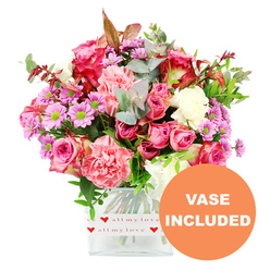 A generous, garden-fresh bouquet fills a clear square glass vase, glowing warmly against a crisp white background that makes every petal and leaf stand out, as if displayed in a bright Epping florist studio near the station. At the heart of the arrangement are plush pink roses, some in deep fuchsia and magenta, others in softer, romantic blush, their layered petals opening in spirals that suggest tenderness and care. Around them cluster ruffled carnations in coral, peach-pink, and gentle pastel hues, their soft frills creating a cloud-like texture. Nestled between these are dainty lavender and purple chrysanthemums, each bloom topped with a lively green centre that adds a zesty, playful energy to the design. Creamy white flowers are scattered throughout, providing a calm counterpoint to the brighter colours so the bouquet never feels overwhelming. Encircling the blossoms is an abundance of foliage: silvery-green eucalyptus with rounded leaves, deeper green sprigs, and a hint of reddish-brown foliage that lends an almost autumnal warmth, reminiscent of walks along Epping Forest's leafy paths. The stems are clearly visible through the transparent vase, which is wrapped at the base with a smooth white band printed with small red hearts and the affectionate phrase "love all my love," subtly suggesting this gift is meant for someone treasured. In the bottom right corner of the image, an orange circular graphic announces "VASE INCLUDED" in bold white capitals, making it easy to imagine this bouquet arriving as a complete, ready-to-enjoy gift for homes around Theydon Bois, Coopersale, or central Epping. The overall feeling is one of thoughtful, uplifting affection, ideal for Valentine's Day, Mother's Day, or simply brightening a loved one's week.