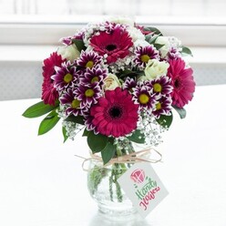 A lush, professionally arranged bouquet of mixed flowers is showcased in a clear, gently textured glass vase, placed against a softly glowing white background that suggests sunlight passing through a window on an Epping side street. The composition is dominated by large magenta gerbera daisies, their petals saturated in deep fuchsia tones and circling velvety dark centres that add drama and focus. Around them cluster bicolour chrysanthemums, each bloom composed of fine petals edged in vivid purple that melt into clean white near the centre, where bright yellow disks give a playful, daisy-like charm. Threaded among these are creamy white spray roses, some just beginning to unfurl, their petals smooth and almost porcelain in contrast to the more textured blossoms. Delicate clouds of white baby's breath float between the larger flowers, offering an airy lightness that softens the intense pinks and purples. Rich green foliage frames the design, with leaves that arch and curl naturally, grounding the colour and making the bouquet feel freshly gathered. The stems are visible through the vase, neatly cut and arranged, the water clear and bright. Around the neck of the vase a piece of rustic twine is tied in a simple bow, from which hangs a small white rectangular tag printed with a stylised pink rose and elegant writing, hinting at a handwritten message inside. The mood is joyful yet refined, perfect for a birthday brunch near Epping Station, a thank-you gift for neighbours in Coopersale, or a bright centrepiece on a dining table after a stroll in Epping Forest, capturing the feeling of celebration and everyday beauty that local florists create for same-day delivery across the area.