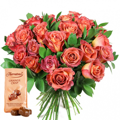 As a florist in the heart of Epping, I see the magic that flowers and fine confections can create-especially when composed so thoughtfully. This image brims with emotional resonance: a magnificent bouquet of open roses radiates from the center, their petals blending warm coral and soft peach with bursts of salmon and deep pink. Some blooms marvel with two-toned or ombre effects: gentle creams and yellows lingering at the heart, then deepening into vivid, rosy-pink washes at each ruffled edge. Lush, dark greenery-fresh as if gathered from the woodland edges of Stonards Hill-frames the round bouquet, inviting the eye to linger and the hand to reach out. In harmonious balance, the Thorntons Orange Crisp chocolates offer gourmet delight. Their elegant pouch, with its signature logo, invites closer inspection, while two glossy, rounded truffles set before it seem almost to glisten under the soft light. The contrast of floral softness and chocolate richness evokes romance, comfort, and joy-a gentle nod to life's sweetest milestones. Whether gifted for Mother's Day, an anniversary, or a spontaneous gesture along Epping High Street, this pairing is an invitation to savor the moment, crafted with the heartfelt artistry and attention to detail that our local customers have cherished for years.