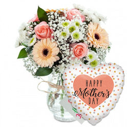 A tender Mother's Day scene unfolds against a clean white backdrop, where every stem and petal has space to breathe, much like the calm early mornings over Epping Forest. In the centre, a clear glass vase, shaped like a classic storage jar, holds a rounded bouquet arranged with the quiet confidence of an experienced local florist. Around the neck of the vase, a length of pale twine is tied in a relaxed bow, lending a touch of rustic charm that feels at home in Epping's mix of character cottages and modern houses. The flowers themselves are a gentle celebration of pastel colour: peach-pink roses fully open, their spiral hearts unfurled into soft, velvety layers, sit alongside large peach gerbera daisies with dark, almost chocolate-brown centres that draw the eye. Airy clusters of white baby's breath thread between the larger blooms, their tiny blossoms forming a soft halo, while compact white chrysanthemums with pale green centres add fullness and a crisp, garden-fresh brightness. Deep green foliage cradles the arrangement, creating contrast and a sense of lively growth, as if the bouquet had just been gathered from a well-tended Epping garden after a spring shower. Behind the flowers, a small, nearly hidden greeting card peeks out in white with hints of colourful lettering. In front, slightly to the right, floats a heart-shaped foil balloon: its white surface scattered with shiny gold and silver spots, framing a peach-coloured heart stamped with the words "HAPPY Mother's DAY" in bold black lettering and elegant script. The overall mood is one of affection and gratitude, perfectly suited for delivering love to mums across Epping, Loughton, and the surrounding area.
