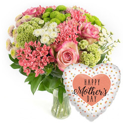 A lavish Mother's Day bouquet with balloon is displayed against a bright white background, the kind of product image you might see when ordering flowers in Epping. The arrangement is gathered into a clear glass vase, allowing you to see the fresh green stems resting in clear water, neatly cut and carefully placed. Above the vase, the flowers swell into a rounded, generous dome of colour. Soft pink roses with delicately darker edges take the lead, their layered petals opening in spirals that feel both romantic and reassuring. Around them, numerous clusters of coral-pink Kalanchoe-like blossoms and other small, spiky florets nestle closely, creating pockets of rich, warm colour. Lime-green button chrysanthemums provide a striking contrast; their compact, textured heads bring a playful, almost mossy look that enlivens the design, reminiscent of the vibrant greens seen around Stonards Hill or Swaines Green in spring. White daisy-style spray chrysanthemums are dotted throughout, their crisp petals and sunny centres catching the light and adding brightness from every angle. Deep, glossy foliage frames the whole display, with leaves peeking between the blooms to give structure and a natural rhythm. To the lower right, a gleaming heart-shaped foil balloon completes the gift. Its light background is sprinkled with irregular metallic gold and soft silver-peach dots, like festive confetti frozen in mid-air. At its centre, a solid peach heart showcases the words "HAPPY Mother's DAY" in an elegant black font, combining bold and script lettering. Together, the fresh flowers and joyful balloon create the impression of a heartfelt Mother's Day surprise, perfect for bringing a burst of colour into a family home anywhere across Epping and the surrounding lanes.