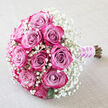 Diagonal on a pale, linen-like background, this bouquet appears as if it has just been laid down by a bride pausing for breath before a ceremony in Epping. The composition centres on plump, multi-toned roses, about a dozen in number, whose petals transition from muted lavender and dusty mauve in the core to saturated fuchsia and deep magenta along the edges. Each rose is carefully placed so that its swirling petals are visible, some blooms tight and domed, others slightly unfurled, creating a rhythmic play of texture and depth. They are enveloped in a cloud of white baby's breath, the countless tiny blossoms of gypsophila forming a soft, lace-like surround. This white halo not only brightens the pink tones but also adds a sense of airiness, like a veil of mist over the roses. A few dark green leaves appear at the edges, hinting at freshness and the natural structure beneath the design. The stems are tied at the lower right with a satiny lilac ribbon that gleams against the neutral fabric, suggesting the polished touch of an experienced local florist. A delicate strand of small pearls spirals over the ribbon wrap, evoking classic bridal jewellery and the quiet refinement of a traditional church wedding, perhaps on the High Street or near the edge of Epping Forest. The photograph captures a bouquet that feels both timeless and contemporary, ideal for celebrating love, commitment, and milestone moments across Epping and nearby villages.