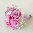 Arranged on a pale, lightly woven beige backdrop, this small bouquet feels like a glimpse of a wedding morning in Epping, captured just before guests arrive. Several full-headed roses form the heart of the design, each one a study in colour: cool lavender and lilac centres gently flow outward into vivid magenta and deep fuchsia rims, so that every petal seems brushed with two tones at once. Nestled intimately around these roses are generous sprigs of baby's breath, their tiny white blossoms clustered in soft, cloud-like tufts that bring brightness and airiness to the compact arrangement. The combination of pastel purples, bold pink edges, and crisp white gypsophila creates a gentle, romantic contrast against the neutral fabric, much like a bride's bouquet standing out against a classic ivory gown. The stems are neatly bound in a rich purple satin ribbon, the material smooth and slightly lustrous, and this handle is meticulously dressed with a spiral of gleaming white faux pearls that wind from top to bottom, giving the bouquet a refined, almost heirloom quality. Positioned diagonally with the handle pointing to the right, the bouquet looks ready to be lifted for a photograph outside Epping Station or carried down the aisle at a local registry office. The scene suggests softness and anticipation: you can almost imagine the faint, sweet fragrance of the roses mingling with the freshness of baby's breath, a hand-crafted design created for a bridesmaid, prom date, or special celebration arranged with care by a nearby florist who understands the area's understated elegance.