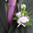 This boutonnière, crafted with mindful attention and local sensibility, nestles against the deep charcoal of a fitted suit and announces itself with calm confidence. The focal white lisianthus, edged in a lively purple ruffle, looks almost luminous, its soft, silk-like petals slightly cupped as if to hold a breath. Surrounding greenery - slender blades, ornamental grass loops, and glossy dark leaves - frames the bloom with a sense of movement and fresh texture, while tiny sprays of gypsophila lend an airy, dewy softness. An upright stem of indigo veronica provides a smart vertical note, balancing the rounded lisianthus and hinting at a refined asymmetry. Behind the main flower, three pale lisianthus buds rise like small promises, suggesting mornings in the garden and new beginnings. The bouquet is tied with a crisp white ribbon whose neat finish reads as both modest and deliberate against the suit lapel; placed next to a magenta tie, the composition becomes an intentional play of colour-romantic but assured. As a local florist serving Epping and its neighbourhoods from near the High Street to the edges of Epping Forest, Flowers Epping composes these details with a sensitivity to texture, scent and season. A faint, fresh fragrance of greenery and bloom clings to the piece, steadying the wearer over hours of celebration. It's ideal for a groom, best man or an honoured guest at a wedding or anniversary - a small work of floristry that carries warmth, craft and the quiet pride of local hands.