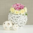 This image shows a beautifully composed floral centerpiece that captures a balance of romance and contemporary style, suitable for a wedding breakfast in Epping or a refined dinner party at home. The focal point is a compact, domed cluster of roses, carefully arranged so that no stem is visible, only a cushion of petals. Around a dozen blooms are used, in a thoughtfully balanced mix of colours: soft, blush-pink roses with a slight lavender tint paired with creamy yellow-ivory roses that lighten the overall feel. The roses are at varying stages of opening, which adds depth and movement, from buds tightly wrapped to more open flowers revealing layered whorls of petals. Interwoven among them are slender sprigs of white baby's breath, their minute blossoms giving a misty, almost bridal veil effect that softens the transition between pink and cream. The bouquet is set into an eye-catching spherical vase made of thin, white-painted wicker or intertwined branches, creating a delicate open lattice that feels both rustic and modern. This globe-shaped container is placed on a crisp white surface that reflects some of the light, brightening the composition. To the left sit two decorative woven balls made from pale natural wood or rattan, one medium-sized and one smaller, their warm tones and open weave echoing the structure of the vase and tying the scene together. The backdrop is a lightly textured beige wall, evenly lit so that the pastel hues and white elements stand out clearly. The overall impression is of a soft, inviting arrangement, ideal for a bridal table, intimate engagement celebration, or a thoughtful floral delivery to an address near Epping Forest or the surrounding residential streets.