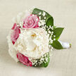 Displayed on a pale, textured fabric that resembles natural linen, this hand-crafted bouquet feels like a quiet breath before the ceremony begins at an Epping chapel or country house. A large, creamy white peony sits proudly at the centre, its petals layered in soft, feathery ruffles that create a plush, rounded focal point. Around it, fuchsia-pink and soft blush roses nestle in close harmony, their tight, spiralled hearts suggesting youthful romance and tenderness. Wisps of baby's breath lace through the arrangement, their tiny white blossoms forming a light veil between the larger flowers and adding an airy, cloud-like halo. Encircling everything, broad, deep green leaves curve and tuck around the blooms, giving a polished outline that contrasts beautifully with the pastel tones. The stems are carefully straightened and wrapped in a sleek white satin ribbon, forming a neat handle that speaks of expert floristry and careful preparation. Gentle, diffused light brushes across the bouquet, catching the sheen on the leaves and the subtle shadows in each petal fold, so that the flowers appear freshly gathered and full of life. Resting on the neutral backdrop, the colours remain soft yet confident, evoking the calm of an early walk along Epping High Street or a peaceful moment under the trees of Epping Forest. This bouquet conveys elegance, sincerity, and the quiet joy of a wedding day, ideal for brides who want a classic, romantic look with a natural, local touch.