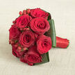 On a warm, light beige fabric that resembles natural linen laid out in an Epping cottage, a compact hand-tied bouquet of red roses lies gently on its side, inviting close inspection. The roses are densely packed into a classic rounded shape, each bloom showcasing velvety petals that curl and unfurl in rich layers, transitioning from vivid scarlet at the centre to deeper crimson and burgundy at the outer edges. Nestled between the blooms are bright red hypericum berries, small and spherical, their glossy skins reflecting the soft light and providing a playful contrast in both texture and form. Surrounding the rose heads, broad dark green leaves have been expertly folded and overlapped to create a clean, modern collar, their smooth, almost satiny surfaces catching subtle highlights. The stems extend to the right and are tightly wrapped in a bold red satin ribbon, the neat finish hinting at the careful, steady hands of a seasoned local florist preparing wedding flowers for a ceremony near St John's Church or a celebration close to Epping Forest. The neutral, warm-toned background allows the bouquet's sumptuous reds and lush greens to take centre stage, while the slightly elevated viewpoint reveals the intricacy of every placement and the balanced symmetry of the design. The arrangement exudes an air of timeless romance and sophistication, equally fitting for a Valentine's Day surprise delivered across Epping, a milestone anniversary, or a bridal bouquet intended to leave a lasting impression in photographs and memories alike.
