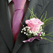 Pinned carefully to a charcoal suit jacket, this blush-pink rose boutonniere tells a story of elegance and tenderness, the sort of detail you might see as a groom waits quietly outside Epping's registry office or a country hotel near Theydon Bois. The central rose is full yet refined, its petals layered tightly at the heart and loosening towards the edges, where they catch the light with a soft, satiny gleam. Around it, delicate stems of gypsophila weave in and out, their tiny white blooms forming a misty halo that softens the strong lines of the lapel. Wisps of slender greenery arc gently away from the rose, adding a fresh, natural rhythm and a hint of garden charm, as though the flowers had just been gathered from a well-kept plot near Epping Forest. The florist has tied the base with a slim pink satin ribbon, its gentle sheen echoing the romantic palette without overwhelming it, and the knot is neatly finished, a subtle sign of experienced, careful hands. Light falls across the arrangement in a flattering way, deepening the charcoal of the suit and making the rose glow softly at the centre. You can almost imagine the cool fabric beneath your fingertips, the faint fragrance of the rose mingling with crisp morning air. This boutonniere is not flashy or overdone; instead, it speaks of quiet confidence, of love expressed through small, meaningful details, and of a moment in time that will be carried through photographs and memories long after the celebrations in and around Epping have ended.