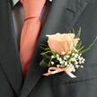 This groom's boutonniere, crafted with attentive hands, presents a single peach rose as its heart - petals curl inward like the first light of dawn, their creamy edges shading into a warm coral-pink that softly mirrors the silk tie at its side. Surrounding the rose, delicate sprays of baby's breath float like tiny clouds, their white clusters lending an innocent, confetti-like quality to the piece. Slim, fresh green foliage threads through the composition, offering graceful lines and a dewy-textured backdrop that lifts the central bloom. The whole is anchored by a low satin ribbon bow, its sheen catching the light and tying the palette together with understated elegance. Against the charcoal suit, the colours read as joyful and sophisticated, each element sharpened by the dark fabric so that petals, ribbon and leaf stand out with intentional clarity. You can almost sense a faint fragrance - a soft, sweet rose note tempered by green, leafy brightness - the kind of scent that feels familiar on a warm spring morning. As a local florist who often dresses wedding parties around Station Road and the market on Epping High Street, I picture this pinned beneath boutonniere pins, steady and secure, a small promise of celebration. Flowers Epping prepares details like this to complement personal style - a gentle, tactile accent that holds meaning and memory for the day.