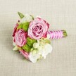 A small, romantic hand-tied bouquet rests horizontally on a light tan, textured linen-style cloth, the neutral background allowing every flower to stand out with clarity. Two fully opened blush-pink roses form the heart of the design, their petals spiralling in tight, layered whorls that deepen to a soft magenta along the outer edges, creating a gentle ombré effect. Around them, bright apple-green button chrysanthemums form neat, spherical tufts, each one covered in tiny, closely packed florets that add a playful, pom-pom texture and a fresh, springlike feel. Creamy white filler blooms, likely petite lisianthus or waxflower, weave between the pink and green, their soft petals and tiny yellow-tinted centres adding lightness and air to the arrangement. Subtle touches of darker green foliage peek out from beneath the blossoms, giving natural depth without overpowering the colour palette. The short stems are neatly trimmed and gathered into a compact handle, tightly wrapped in smooth, luminous fuchsia satin ribbon. Over the ribbon, a fine strand of white faux pearls is carefully crisscrossed, catching the soft, even light and lending a bridal sense of polish and grace. The overall look is elegant yet fresh, as if prepared for a bridesmaid walking through a ceremony at a country house near Epping Forest or a registry office just off Epping High Street. It feels intimate, crafted for the hand that holds it and the camera that captures it, a delicate posy suited to weddings, engagement celebrations, or a special dinner at home in Epping and the surrounding villages.