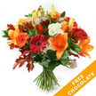 Against a pristine white background, a hand-tied bouquet stands upright like a small celebration of autumn light. Large orange lilies with trumpet-shaped blooms and visible stamens take centre stage, their waxy petals catching a soft, studio glow. Around them, ruby-red and bright-orange Gerbera daisies smile with dense, circular petals, while clusters of creamy white Lisianthus with ruffled edges add a crisp, almost cool contrast. Small, star-like Astrantia with purple-pink centres peek through, their intricate heads lending a delicate, lacy texture against the bolder blooms. Deep green leaves-likely ruscus or salal-form a glossy, verdant base, and the composition is punctuated by deeply lobed oak leaves in burnt orange and reddish-brown that immediately read as autumn foliage. The stems are neatly gathered and secured with a dark twine or raffia, the knot visible and slightly rustic, affirming the traditional hand-tied style.
The bouquet feels tactile and fragrant: the smooth, satiny petals of the lilies, the firm, velvety discs of the gerberas, and the papery crispness of the oak leaves combine into a warm, seasonal scent you can almost imagine. This arrangement is ideal for saying thank you, marking a birthday in late October, or bringing autumn cheer to a home on Epping High Street or after a walk through Epping Forest. In the lower right corner of the image a bright orange diagonal banner overlays the scene, boldly reading FREE CHOCOLATE in white capital letters-an eye-catching promotional detail included with the product image. The overall effect is expertly composed, warm, and welcoming, evoking a crisp, golden autumn afternoon in Epping.
