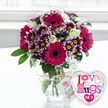 On a clean, pale surface that recalls a bright kitchen or hallway in Epping, a lush floral arrangement takes centre stage in a classic rounded glass vase. The bouquet bursts with colour, anchored by several large gerbera daisies in a saturated magenta-fuchsia hue, their smooth petals radiating from dark, velvety centres that lend the design a dramatic core. Nestled among them are eye-catching chrysanthemums, each bloom displaying a ring of deep purple petals that gently fade to crisp white at the tips, framing cheerful yellow centres that seem to glow. Creamy white roses, with petals that curl softly at the edges, add a romantic, timeless touch and balance the bolder tones. Fine, airy sprays of white baby's breath weave between the larger flowers, creating a light, cloud-like texture that softens the arrangement and adds depth. Fresh green leaves peek out around the edges, giving the bouquet a full, rounded form that feels both carefully designed and naturally abundant. The clear glass vase reveals straight, neatly trimmed stems immersed in water, and at the neck, a length of rustic natural twine is tied into a simple bow. From this twine hangs a small white gift tag, adorned with a delicate pink rose illustration and handwritten-style green lettering that subtly hints these are hand-chosen blooms from a caring florist. Positioned just to the right of the vase is a shiny, heart-shaped foil balloon with a reflective silver border and white centre, decorated with playful, multi-coloured lettering spelling Love n' Hugs 24-7, underlined with tiny hearts. The softly blurred, bright background suggests daylight streaming through a nearby window, creating an uplifting scene that suits romantic surprises, birthday treats, or thoughtful same-day flower delivery around Epping.