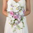 The image shows a bride from the waist down, her white strapless gown forming a smooth, luminous canvas for a sumptuous cascading bouquet that tumbles elegantly towards the floor. At the heart of the arrangement, wide-petalled white Phalaenopsis orchids unfurl in generous arcs, their creamy surfaces catching the light and their yellow-orange centres adding a subtle, sunlit glow. Around and between the orchids, layers of pink and lavender-toned roses cluster tightly, some with classic, spiralled petals and others with the looser, romantic form of garden roses in gentle dusty blush. These roses bring a sense of depth and softness, as though each bloom has opened slowly over a cool Epping morning. Nestled amongst them are lisianthus buds and blossoms in white with the faintest tips of lavender, curving gracefully and giving a sense of natural, garden-like movement. Airy sprigs of baby's breath weave through the design, their tiny white flowers creating a veil of lightness around the more substantial forms, while slender stems of pale lavender stock introduce height, texture, and a hint of fragrance you can almost imagine drifting on the air. The bouquet narrows as it descends, forming a classic teardrop silhouette that flatters the line of the dress and feels perfectly suited to ceremonies in Epping's churches or intimate celebrations near the forest. The soft beige background keeps the colours gentle and cohesive, allowing every flower to stand out without distraction, as if the bouquet were being presented for a final, admiring glance before the bride steps out for her walk down the aisle.
