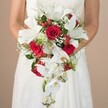 Elegance and artistry converge in this close view of a bride, clad in a white lace wedding dress, delicately holding an opulent cascading bouquet-a floral homage to Epping's tranquil beauty. Set against a neutral, softly blurred background evocative of Epping's quiet lanes, the bouquet comes to life in her gentle embrace. Showcasing the skill of Flowers Epping's local florists, the composition features grand white lilies radiating confidence, their wide petals framing golden stamens at the center. Ribboning below, delicate Phalaenopsis orchids trail like streams flowing through nearby Epping Forest, their intricate forms and subtle yellow accents adding softness and dimension. Red roses, lush and velvety, command attention amid the whites, conveying depth of emotion and classic bridal romance. Airy baby's breath and sprigs of glossy red hypericum berries highlight the bouquet, their fine textures and bright notes contributing a playful yet refined finish. Surrounding foliage-fine green ferns and darker, structured leaves-provides natural volume and visual harmony, with the occasional spiky element suggesting Epping's rich woodland character. The bride's arms cradle the bouquet confidently yet gently, as though presenting a work of art crafted for a day to remember. Ideal for wedding celebrations across Epping and nearby villages, this arrangement captures the essence of love, tradition, and a touch of modern flair, welcoming all who pause to admire the timeless beauty of a local wedding day.