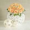 A refined floral centerpiece sits proudly on a bright white table, composed of a generous, domed cluster of soft peach and apricot roses, each bloom tightly cupped with deeper orange hearts and paler, almost blush-toned outer petals that glow as if lit from within. Nestled between the roses are airy sprays of tiny white baby's breath, their cloud-like blossoms softening the arrangement and adding a gentle, romantic haze. A few deep green leaves peek out from beneath the flowers, grounding the design with a natural contrast that makes the warm tones of the roses feel even more inviting. The bouquet rises from an eye-catching spherical vessel crafted from intricately woven white strands, resembling delicate string or fine rattan crisscrossed into an open lattice that lets you glimpse the stems inside. In front of this main vase, two smaller decorative spheres made from the same white strands rest on the smooth tabletop, one slightly larger and patterned like an abstract, minimalist football, the other more loosely woven and organic in feel. Behind the display, a soft, light beige wall with subtle texture offers a calm, uncluttered backdrop, allowing the intricate weaving and the glowing peach roses to take center stage. The whole scene is evenly lit with soft, natural light, evoking the feeling of a peaceful afternoon in an elegant Epping home near the High Street, ready to welcome guests for a wedding brunch or intimate anniversary dinner. The arrangement suggests quiet celebration and thoughtful design, the kind of modern yet romantic centerpiece that local clients often request for stylish events and home styling across Epping and the surrounding lanes.