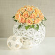 A study in texture and soft colour, this artistic design pairs honeyed apricot roses with airy clouds of white gypsophila, arranged in a perfect, domed embrace above a white rattan sphere. Each rose blooms with layered petals that graduate from creamy peach at the core to deeper apricot at the petal edges, their velvety surfaces reflecting light in a way that feels almost tactile. Baby's breath winds between the heads, its fine stems and tiny blossoms adding a frothy veil that diffuses the composition and lifts the silhouette. The woven sphere beneath functions as both vessel and sculptural base: its organic criss-crossing lines echo the stems' movement and allow glimpses of shadowed space, giving the bouquet a sense of weightless poise. Green leaves, glossy and cool, peek through to provide contrast and to frame the colour palette with fresh, botanical notes. Imagine this piece set on a long table at a reception near the edge of Epping Forest or sitting bright and quiet on a mantel along the High Street-it brings together modern decor sensibilities and timeless floral romance. The fragrance is soft and powdery, a gentle mix of rose and green stem notes reminiscent of a summer morning. As a local florist in Epping I place each bloom with attention to balance and longevity, ensuring the arrangement reads beautifully from every angle and suits occasions from anniversaries to bridal events. For those seeking a refined, tactile bouquet with a contemporary anchor, this design offers a thoughtful, elegant solution.