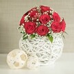 A beautifully balanced floral still life unfolds in this image, dominated by a dome of deep red roses that seem to glow against a calm, neutral backdrop. Between twelve and fifteen roses sit closely together, each bloom open and velvety, with petals that curve inward in tight, elegant spirals, giving the arrangement a polished, almost sculptural quality. Fine sprays of white baby's breath thread their way delicately between the roses, their small blossoms appearing like tiny stars scattered across a crimson sky. The occasional glimpse of fresh green leaves underneath softens the composition further, subtly suggesting the natural woodland greens that edge the Epping area. Cradling the bouquet is a striking, bulbous white vase made from interlaced strands arranged in an open-weave lattice. The crisscrossing white bands create a basket-like effect that feels both contemporary and lovingly handmade, similar to artisanal décor pieces found in the independent shops around Epping High Street. The vase is set on a smooth, pristine white surface, perhaps a dining table prepared for a wedding reception or an anniversary dinner at home. To the left, two decorative spheres made from pale, woven strips echo the same tactile texture, one larger and more prominent, the other tucked just behind, adding a gentle sense of movement and depth. The background is a soft wash of light beige, faintly textured and pleasantly blurred, ensuring that the vibrant reds and intricate weaving remain in focus. Crafted with the eye of a local wedding florist, this arrangement radiates romance, making it a perfect centrepiece or gift for intimate celebrations and same-day deliveries in and around Epping.