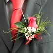 A close, intimate view of a wedding buttonhole captures a single cerise-pink rose bud resting against the charcoal grey lapel of a finely cut suit, as if the wearer is pausing on Epping High Street before walking to the ceremony. The rose is partially opened, its velvety petals elegantly curled inward, creating a rich spiral of fuchsia tones. Encircling the bloom is a soft halo of white gypsophila, each tiny flower crisp and airy, bringing lightness and contrast to the composition. Mixed greenery frames the rose: some leaves are broad and glossy, reflecting a gentle sheen, while slender, feathery sprigs add texture and a sense of natural movement reminiscent of the paths through Epping Forest. At the base of the floral cluster, a vivid red satin ribbon is tied into a neat bow, its luxurious shine matching the bold red silk tie that lies smoothly beneath the lapel. The suit appears to be crafted from a high-quality wool blend, with a subtle pattern on the grey dress shirt that shows just behind the tie's knot. Soft, even lighting reveals every detail, from the fine threads of the fabric to the delicate veins in the foliage. The overall impression is one of quiet romance and refined style, perfect for a groom or groomsman attending a wedding in Epping, where the buttonhole becomes a small but powerful symbol of love, promise, and the joy of a day carefully planned and beautifully shared.
