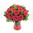 A lavish bouquet of deep crimson red roses stands proudly against a pure white background, each bloom captured at its most beautiful, as if freshly arranged in a local Epping flower studio. The roses are tightly clustered, forming a generous, rounded dome where no stem looks out of place. Every head is perfectly shaped, from the tightly furled central petals that spiral inward like a secret, to the outer petals that gently unfurl and curl back, revealing velvety layers and a rich, saturated ruby tone. Nestled between the blossoms is a collar of dark, glossy green foliage, likely Ruscus or a similar filler leaf, its smooth surfaces catching the light and heightening the intensity of the red. The leaves provide a cool, fresh contrast, framing the roses and adding depth to the arrangement, much like the greenery that lines Epping Forest pathways. All of this beauty is gathered in a translucent ruby-red glass vase with a subtly twisted, spiralled design etched along its sides. The vase flares slightly at the top and settles into a solid, weighty base, giving the composition a sense of poise and permanence on a dining table in a High Street townhouse or a cosy cottage just off Bower Hill. Through the coloured glass, you can see the rose stems, slightly magnified and tinted, reinforcing the impression of abundance. Bright, even lighting reveals every petal edge and leaf vein, suggesting flowers at the peak of freshness, ideal for an anniversary, a Valentine's surprise, or an unforgettable romantic gesture delivered across Epping and the surrounding lanes.