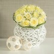 On a smooth white table, a romantic floral display captures the eye with its subtle blend of colour, texture, and light. At the centre, a rounded bouquet of light yellow roses forms a flawless dome, each rose perfectly open so that the petals curl outward in generous layers, giving a plush, cushion-like effect. The gentle yellow hue, somewhere between buttercream and soft primrose, radiates warmth without ever becoming overpowering. Laced between the roses are delicate tufts of white baby's breath, their tiny blossoms creating a fine, cloud-like veil that lightens the mass of petals and adds a feathery contrast. A narrow band of vivid green foliage encircles the base, like a tailored trim that frames the flowers and connects them visually to the vessel. The arrangement is set in a distinctive white woven vase, its structure formed from slender, interlocking strands that create an open, basket-style lattice. The shape is softly curved and slightly cinched at the middle, giving it a graceful, modern profile. Light slips through the gaps in the weave, casting gentle shadows onto the glossy surface below. To one side, two woven spheres made from pale, natural-looking material mirror the texture of the vase, their different sizes adding a balanced, sculptural quality to the composition. The background is a calm, blurred neutral, similar to the soft tones of an Epping boutique or gallery, ensuring complete focus on the flowers. The overall impression is of quiet elegance and joy, perfectly suited to wedding table arrangements, anniversary dinners, or sophisticated gatherings in homes around Epping.
