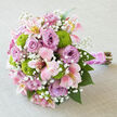 On a pale, textured fabric that resembles light grey or soft stone-washed linen, a lush bridal bouquet lies gently on its side, its rounded shape carefully composed and full of movement. The arrangement is a serene conversation between lavender, blush pink, and bright green, the colours harmonising the way early evening light settles over the lanes of Epping. Large lavender roses, open and luxurious, dominate the bouquet; their layered petals curl and fold like soft silk, creating pools of shadow and light at their centres. Nestled among them, pink and creamy alstroemeria stretch out their star-shaped blooms, each petal patterned with delicate streaks and freckles of deeper pink and subtle golden yellow that lend an almost painterly quality. Popping through the pastels, vibrant chartreuse green button chrysanthemums form tidy, spherical clusters, their texture tight and almost fuzzy, bringing energy and a crisp contrast that feels both joyful and contemporary. Threaded all throughout, tiny sprays of white baby's breath soften every edge, appearing like a dusting of fine lace or a veil of tiny stars scattered between the larger flowers. The bouquet's stems are aligned and wrapped in a wide, glossy pink satin ribbon, smooth and slightly reflective, giving the handle a polished, bridal finish. A slim string of petite white pearls winds around the ribbon, adding a touch of vintage elegance, as if chosen to match heirloom jewellery worn for a ceremony near Epping High Street. The exposed stems at the base introduce a natural, grounded note, with hints of green and woody brown. This bouquet feels romantic yet fresh, perfect for weddings, bridal showers, or special celebrations across Epping and the nearby villages, where understated sophistication and heartfelt beauty are treasured.