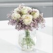 A softly lit, close-up photograph of a refined white and lavender flower arrangement shows a small cluster of creamy white roses opening in gentle spirals, cradled by a cloud of lilac-toned statice. Each rose sits at the heart of the bouquet with petals that appear velvety and thick, their centres carrying the faintest blush of buttery ivory, suggesting flowers cut fresh that morning from a cool flower room. Surrounding them, the statice forms an airy halo of tiny papery blooms, their muted lavender and pale purple shades adding depth without overpowering the roses' calm purity. The bouquet is gathered into a clear glass vase with a distinctive crackled or ice-effect texture; the irregular pattern catches the natural daylight and scatters it in tiny glints across the surface, like frost melting on a window. Through the water, the straight green stems are clearly visible, cleanly trimmed and arranged, reinforcing a sense of care and professional craftsmanship. The vase rests on a bright white tabletop that reflects the light upwards, casting soft, diffused shadows and giving the whole scene a bright, peaceful clarity. The background is blurred into a smooth gradient of pale grey, with no distractions, making the flowers feel like a quiet focal point in a calm Epping home, perhaps on a windowsill looking towards Epping Forest. The mood is one of gentle tranquility and understated elegance, ideal for a thoughtful sympathy gesture, a peaceful bedside arrangement, or a soothing gift delivered to someone in the local area who needs a moment of calm and reassurance.