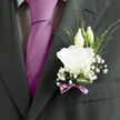 A refined wedding boutonnière rests against the lapel of a well-tailored dark suit, the scene evoking the gentle atmosphere of a ceremony somewhere near Epping Forest or a country house on the edge of town. The focal point is a generous white lisianthus bloom, its petals soft and almost translucent, curling outward in layered, ruffled folds that suggest both innocence and quiet sophistication. The flower seems freshly opened, like a breath of cool air on a spring morning along Epping High Street. Behind it, two slender buds rise gracefully, still tightly closed, their pale green tips hinting at blossoms yet to unfurl-an understated metaphor for new beginnings and shared futures. Surrounding the central bloom, airy clusters of baby's breath weave in and out, each tiny white floret catching the light and creating a halo-like softness around the boutonnière. Fine strands of green foliage slip between the flowers, giving a sense of movement and keeping the design from feeling too rigid or formal. At the base, a single deep green leaf is artfully curled, its smooth, glossy surface providing structure and a natural cradle for the stems. Wrapped around the binding is a slender ribbon in muted mauve, tied into a simple bow that subtly echoes the tone of the groom's tie without overpowering the composition. The combination of textures-silky petals, feathery baby's breath, polished foliage, and the satin ribbon-creates an impression of thoughtful craftsmanship, as if a local Epping florist has taken extra care to ensure this small piece speaks of tenderness, hope, and the quiet joy of saying I do.