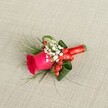 A richly detailed boutonniere is laid carefully on a softly textured, light beige linen background, the weave of the fabric echoing the rustic elegance of a countryside celebration in Epping. At its heart sits a single, deep fuchsia rose, petals tightly cupped at the centre and gradually unfurling into a velvety spiral of colour, suggesting both fresh fragrance and romantic anticipation. Around the rose, tiny white blossoms of baby's breath gather in airy clusters, their cloud-like softness creating a gentle halo that lightens the design. Threaded between these delicate blooms, small, glossy red hypericum berries catch the light, adding a festive sparkle and a subtle hint of seasonal warmth. Wisps of fine fern-like greenery extend gracefully from the sides, while broader, dark green leaves are folded and curled with a practiced florist's touch, giving shape, depth, and a polished outline. The stems are neatly bound with a narrow band of bright red satin ribbon, tied firmly and finished with a small, tidy knot that glows against the neutral fabric. Viewed from a slightly elevated angle, every layer of texture and colour is visible, from the sheen of the berries to the matte softness of the petals. It feels like the perfect lapel flower for a groom or groomsman walking through Epping High Street to a wedding at a local venue, a compact emblem of joy, love, and careful craftsmanship that would suit formal events, proms, or any special occasion where elegance and sentiment matter.