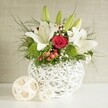 A graceful floral centerpiece is presented in calm, diffused light, as if set on a wedding table overlooking the greenery on the edge of Epping Forest. At the center of the scene stands a spherical white vase, its structure made from interlaced strands that curve and intersect to form an elegant open-weave pattern, giving it a contemporary yet organic feel reminiscent of artisan pieces found in small shops along Epping High Street. Emerging from this sculptural base is a generous bouquet of white lilies, red roses, and textured greenery. Several lilies are fully open, their wide, silky petals creating star-like forms with prominent golden stamens, while a handful of green buds rise above them, suggesting the arrangement will continue to evolve over the coming days. Nestled among the lilies, a rich red rose draws the eye with its velvety, tightly layered petals, bringing a focal point of deep, romantic colour that suits both weddings and intimate dinners at home. Bright green pom-pom blooms add cheerful, rounded forms and a fresh, almost moss-like texture, contrasting beautifully with the smooth lily petals. Sprays of tiny white baby's breath weave delicately through the design, softening the transitions between blooms, while clusters of small red berries tuck into the greenery, adding a subtle festive note perfect for celebrations in and around Epping. Airy, fern-like fronds and broader dark green leaves spill gently over the rim of the vase, giving the arrangement a relaxed, garden-inspired silhouette. To the left, two decorative balls in pale woven material echo the vase's pattern and introduce a sense of balance. The neutral background allows every colour and texture to stand out, capturing a moment of quiet, refined beauty ideal for a wedding table arrangement or special event centerpiece.